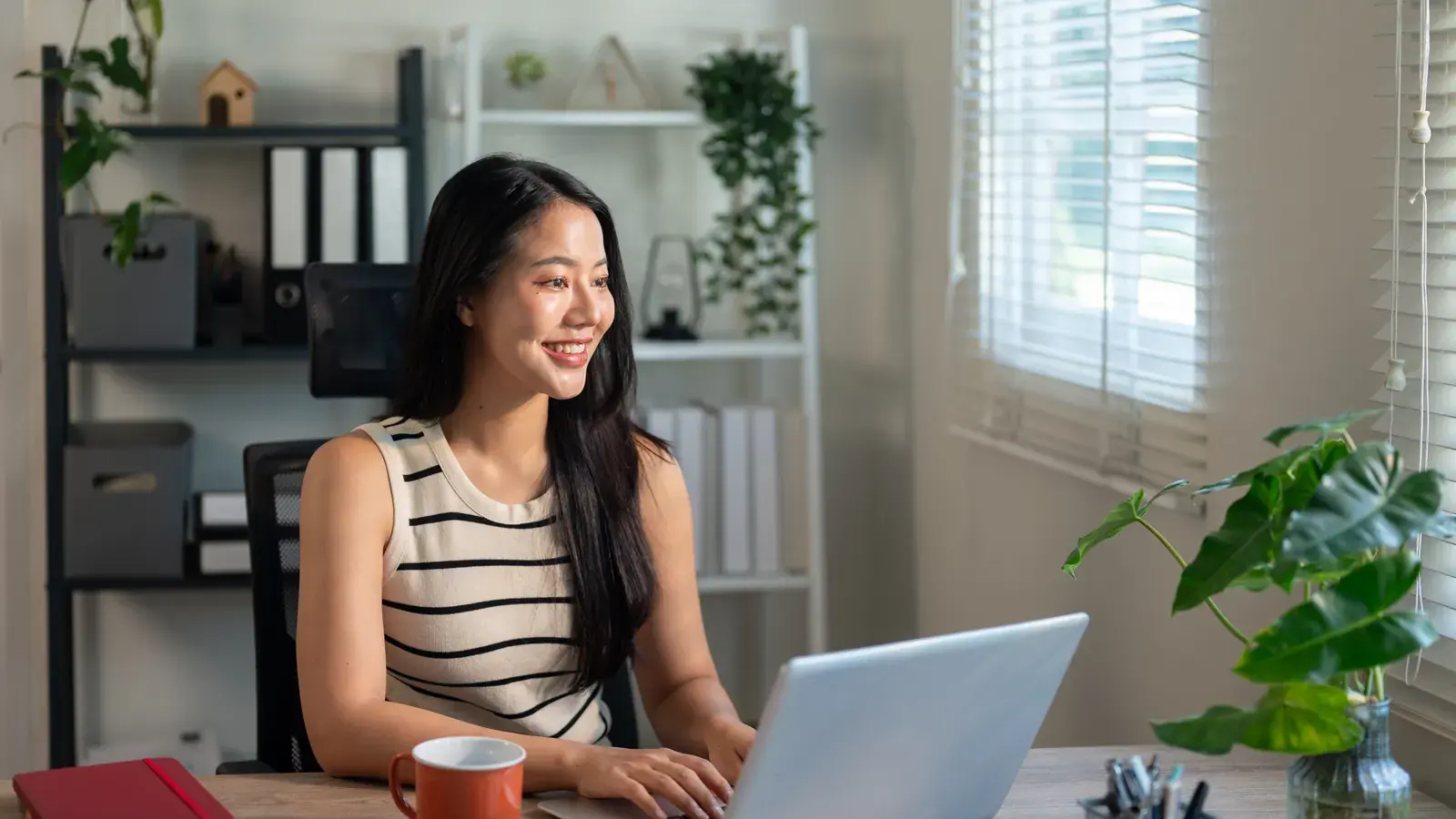 An Indonesian remote worker at a home desk, working on a laptop in warm daylight.