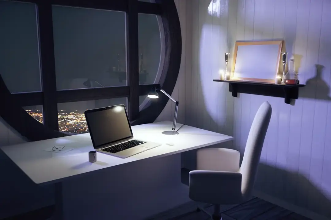 A desk lamp and laptop at night, city lights outside the window.